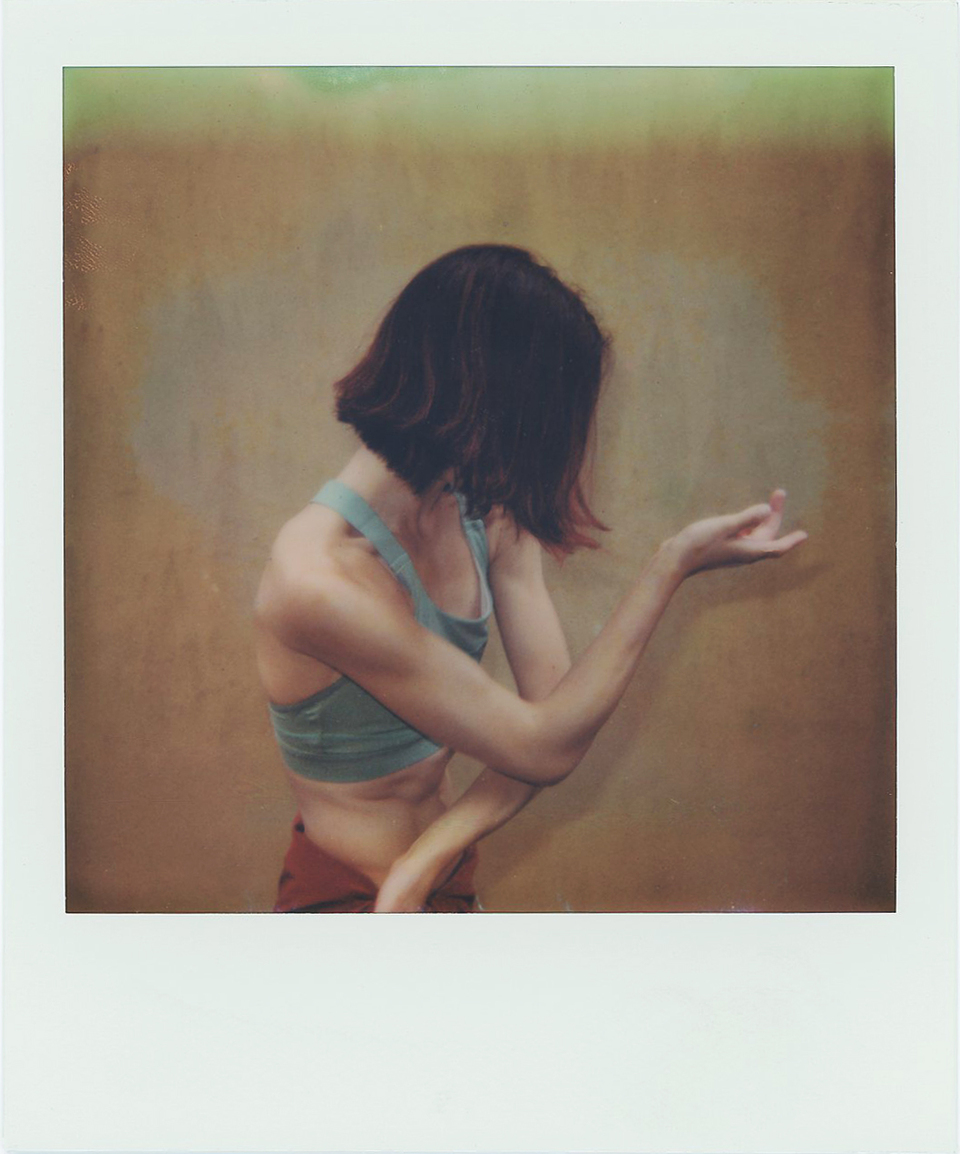 A person with short brown hair, wearing a teal crop top, stands against a muted brown background. They turn their head away from the camera and bend one arm upward, hand at chin level, in a thoughtful or expressive pose.